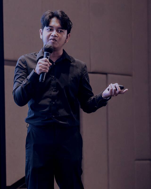 Eryawan Presma Yulianrifat speaking to a seated room, a handheld microphone in his right hand and his left hand open mid-explanation, with a presentation slide in Indonesian lit up at the edge of the frame.